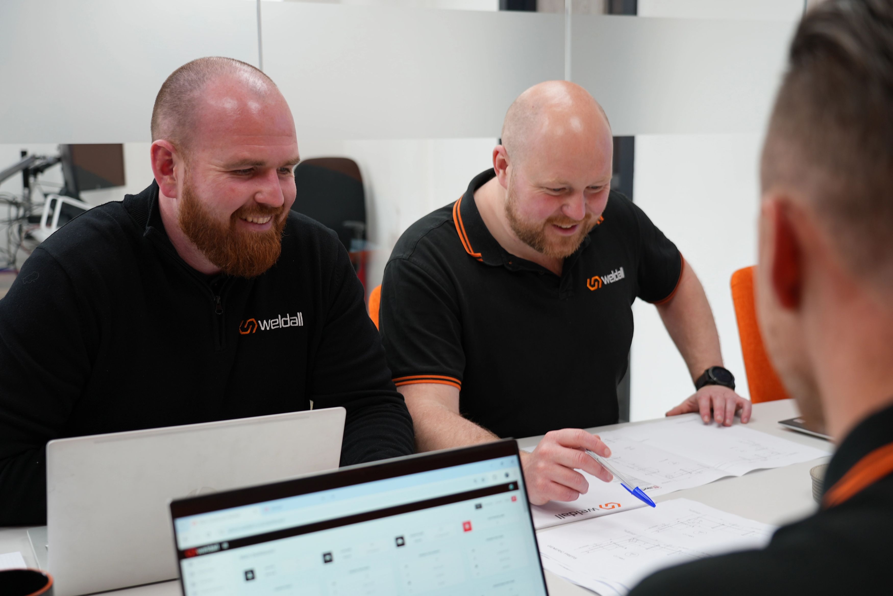 Weldall team members reviewing engineering drawings and laptop screens during an office meeting.
