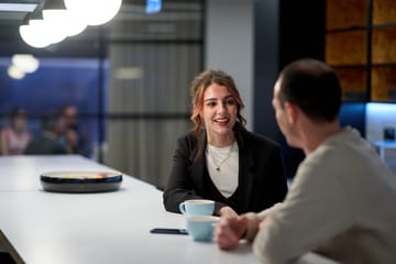 Emma and Andy having a casual conversation over coffee in a modern office lounge