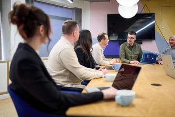 Emma, Ryan, Andy and Jamie in a team meeting around a conference table with laptops and coffee mugs.