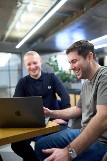 Nick and Jacob smiling while working together on a laptop in a modern office.